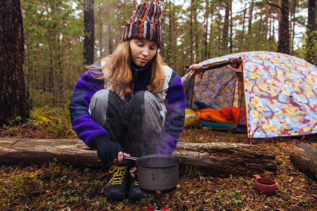Nuori nainen istuu metsässä retkitulen äärellä, keittää ruokaa kattilassa. Taustalla värikäs teltta ja varu...